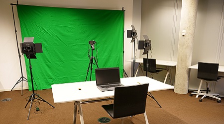 A green screen is set up with lights and a tripod in front of it, as well as a laptop sitting on a table with a chair