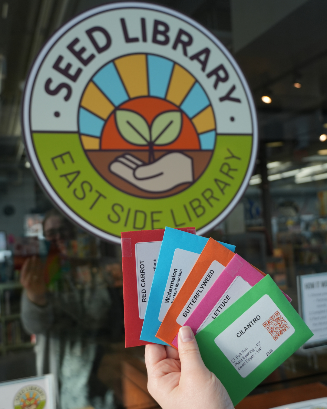 Seed Library East Side Library with a hand holding multicolored seed packets for Cilantro, Lettuce, Butterfllyweed, Watermelon, and Red Carrot