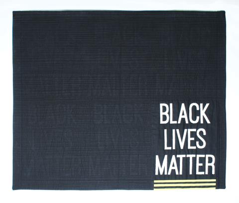 art quilt by Toni Corbett with a black background and the words "Black lives matter" in white in one corner and quilted into the surface five other times