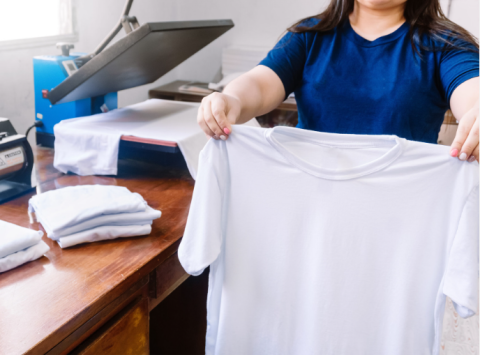 Woman holding a white t-shirt, a heat press in the background on a wooden table