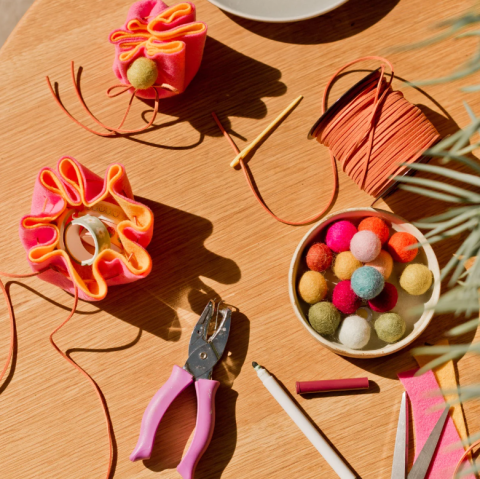 Overhead view of a brown table with a hole punch, needle and thread, puff balls and pouches laid on it